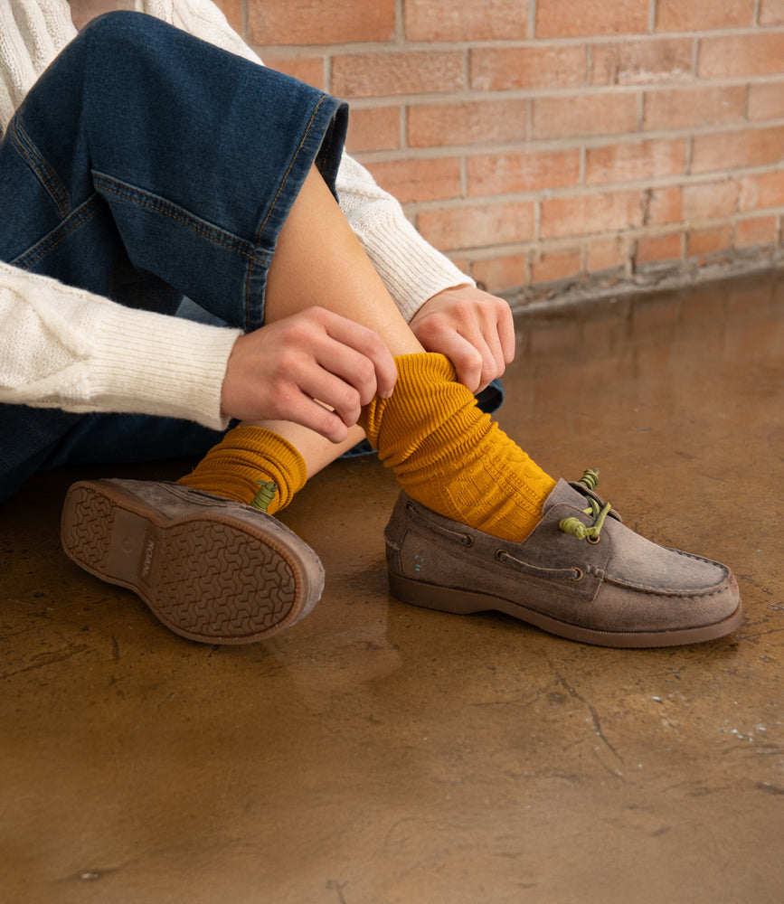 A person sits on a polished floor in blue jeans and a cream sweater, adjusting mustard yellow socks with Roan’s Abide brown women’s leather boat shoe. A brick wall is in the background.