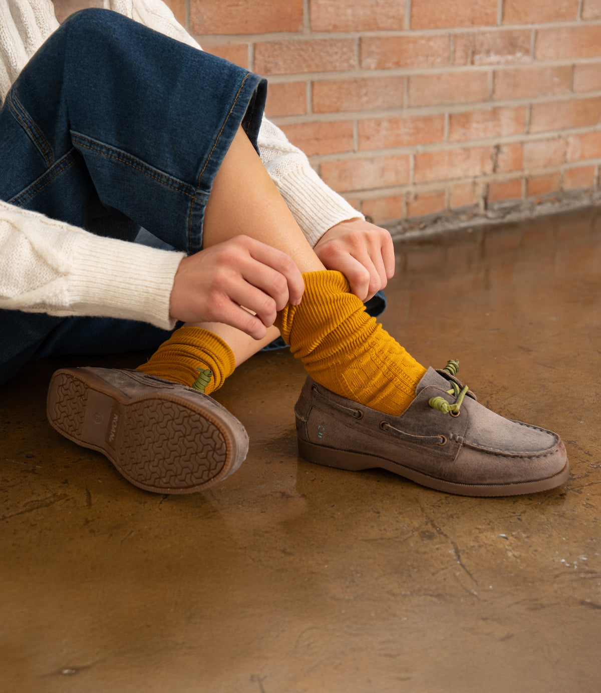 A person sits on a polished floor in blue jeans and a cream sweater, adjusting mustard yellow socks with Roan’s Abide brown women’s leather boat shoe. A brick wall is in the background.