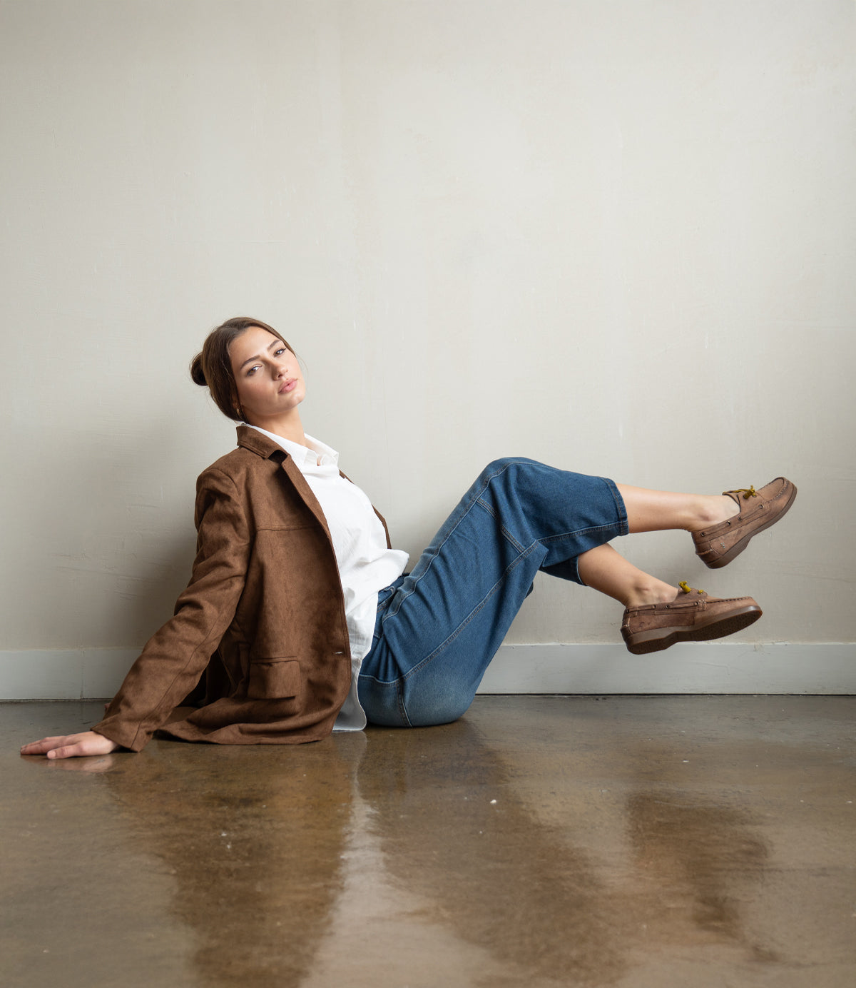 
                  
                    A woman in a brown blazer, white shirt, and blue jeans sits on the floor against a plain wall, confidently showcasing Roan's Abide women’s leather boat shoes—durable and water-repellent—as she looks toward the camera.
                  
                