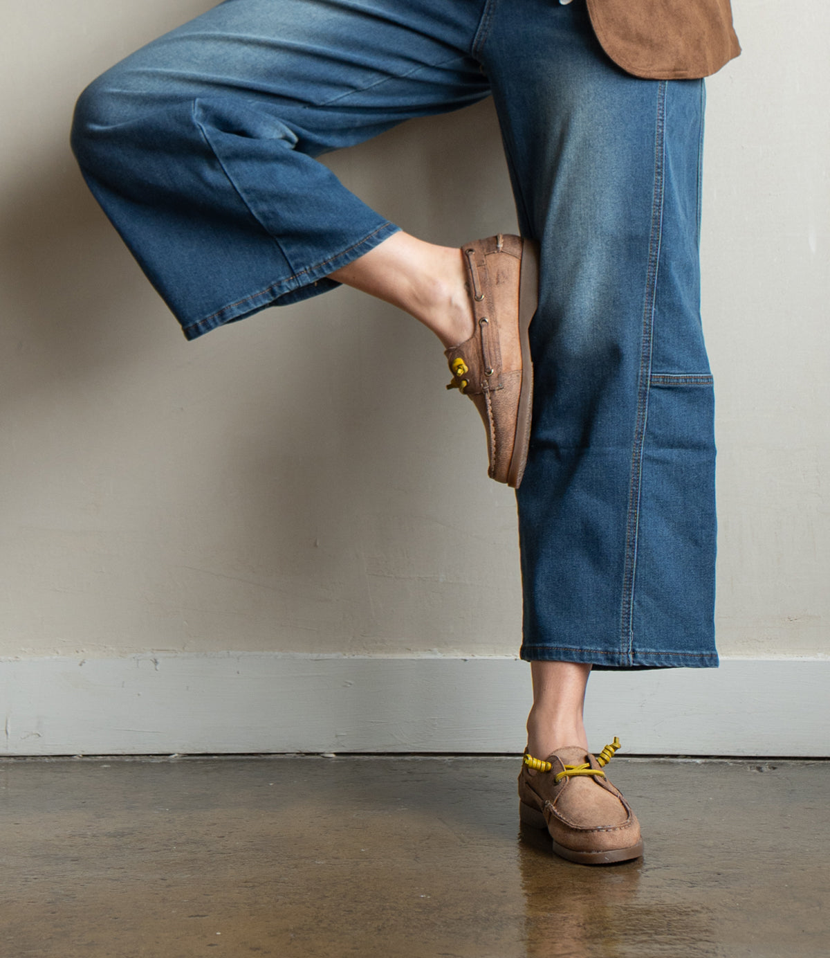 
                  
                    A person wearing blue jeans and Roan's Abide women's leather boat shoe with yellow accents stands on a polished concrete floor, one leg bent with their foot against the wall.
                  
                