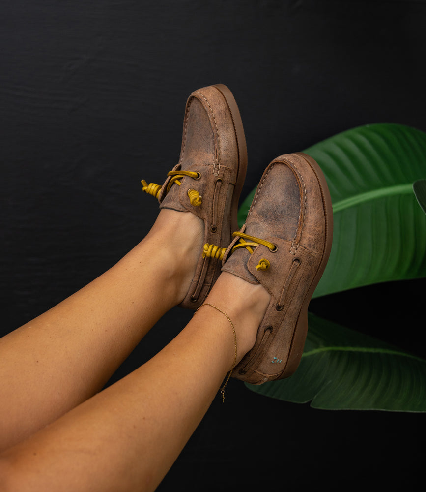 
                  
                    A pair of Roan Abide women's leather boat shoes in brown with yellow laces, worn and poised above green leaves on a dark background—timeless elegance in every step.
                  
                