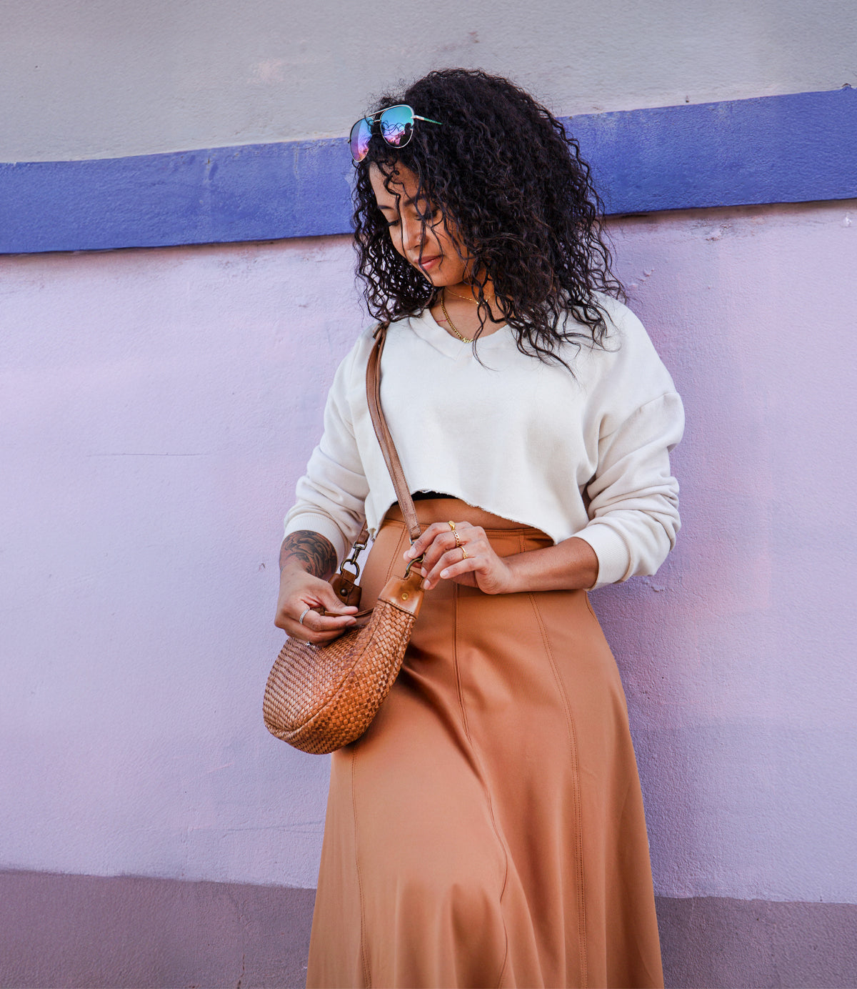 A woman with curly hair, sunglasses on her head, a white sweater, and a brown skirt stands by a pink and purple wall, holding the Roan Agenda lambskin leather crossbody clutch.