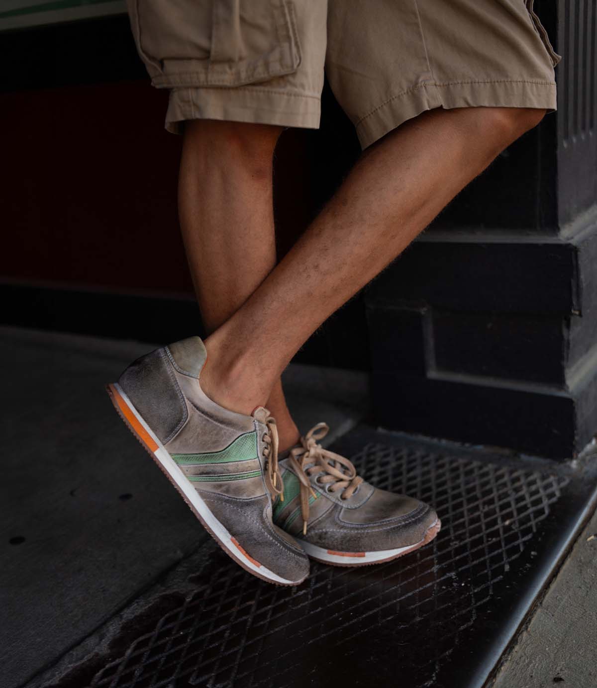 
                  
                    A person in tan shorts stands with one leg crossed, wearing Roan Alatar sneakers in worn gray leather and suede with green and orange accents.
                  
                