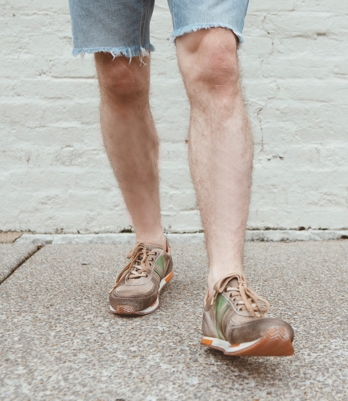 A person wearing frayed denim shorts and Roan Alatar retro-inspired leather sneakers with contrasting green stripes walks on a textured surface against a white brick wall background.