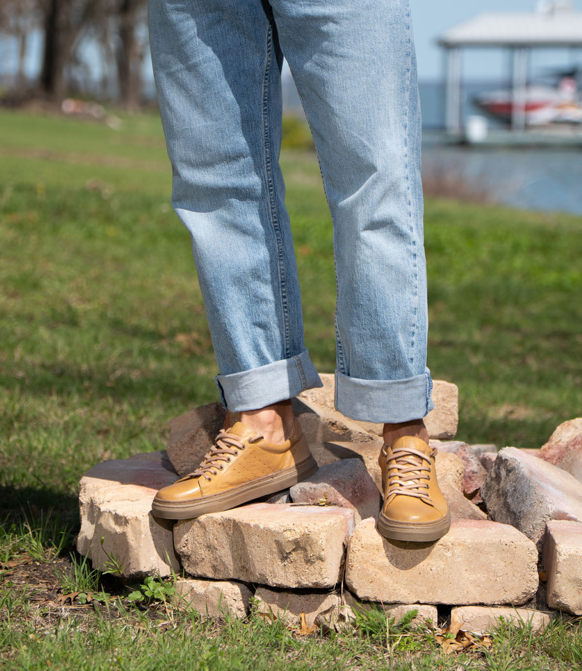 A person stands outdoors on light-colored bricks, wearing Roan's Albright tan leather sneakers with a cushioned footbed and cuffed blue jeans, with grass and a lake in the background.
