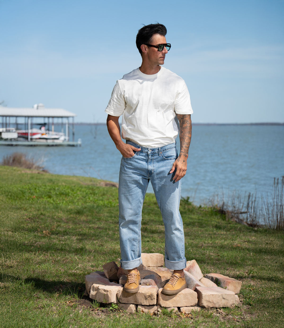 A man in a white T-shirt, light blue jeans, and Roan's Albright tan boots with a cushioned footbed stands on a stone circle near a lake, with grass and a dock in the background.