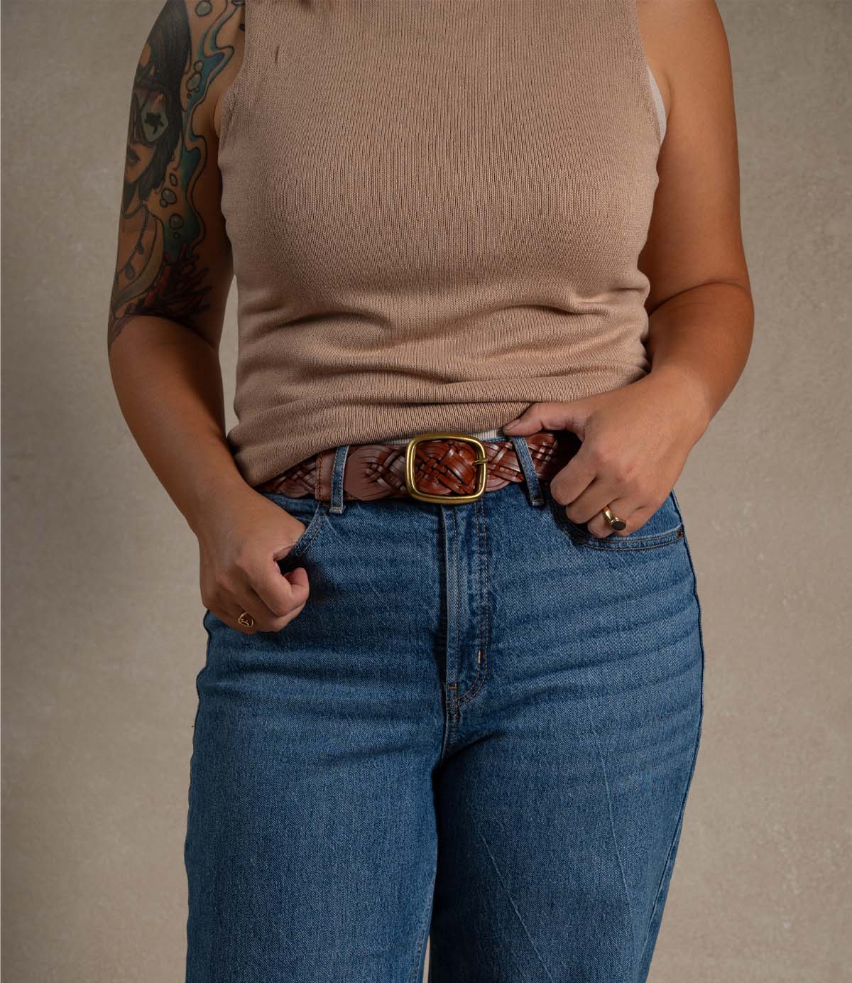 
                  
                    A person wearing the Alder sleeveless beige top by Roan, paired with blue jeans and a braided leather belt with a rounded metal buckle, displays a tattooed arm while standing against a plain background.
                  
                
