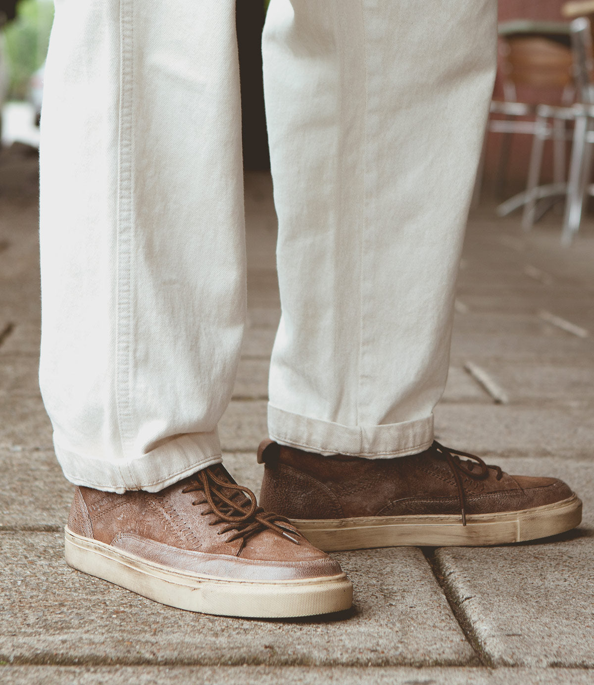 Person wearing white pants and brown Roan Anduril leather shoes with laces, standing on a paved surface, embodying a casual daily wear style.