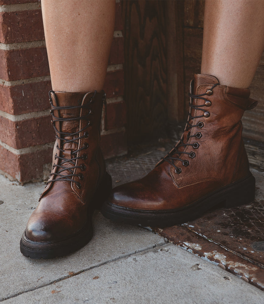 A person is standing on a concrete floor near a brick wall, wearing Roan Awol brown leather lace-up boots with rope laces.