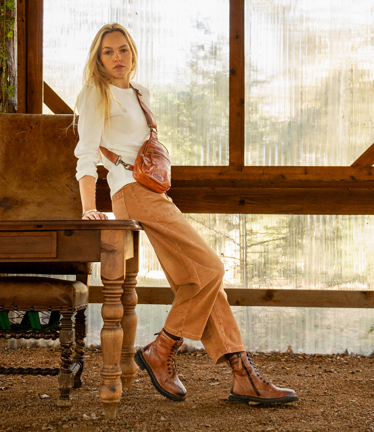 
                  
                    A woman in casual attire and boots, carrying the Roan Trickle compact leather bag, leans against a wooden table in a rustic room with large windows framing forest views.
                  
                