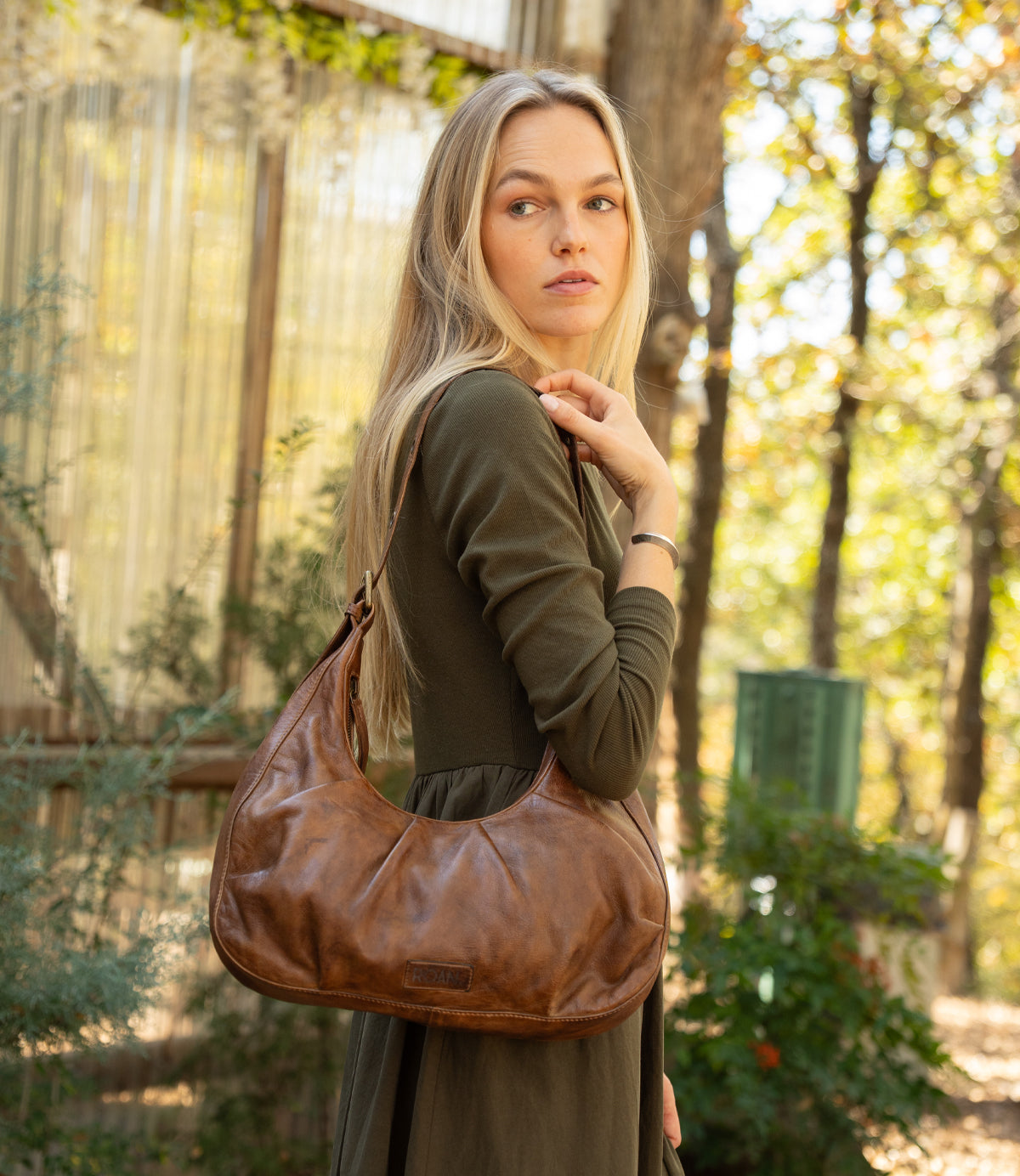 A woman in an olive green dress stands outdoors, holding the Roan "Behalf" crescent silhouette bag with an adjustable buckle strap. Trees and rustic structures appear in the background.