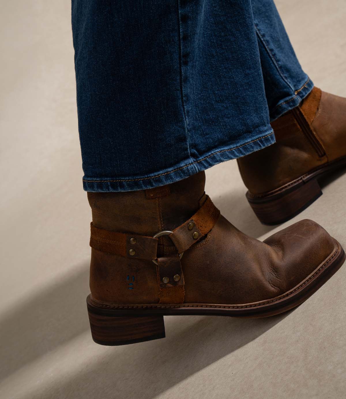 
                  
                    A person wearing blue jeans and Roan’s Bonsai boots—brown, full-grain leather harness strap boots featuring a thick wooden heel—stands on a light-colored surface.
                  
                