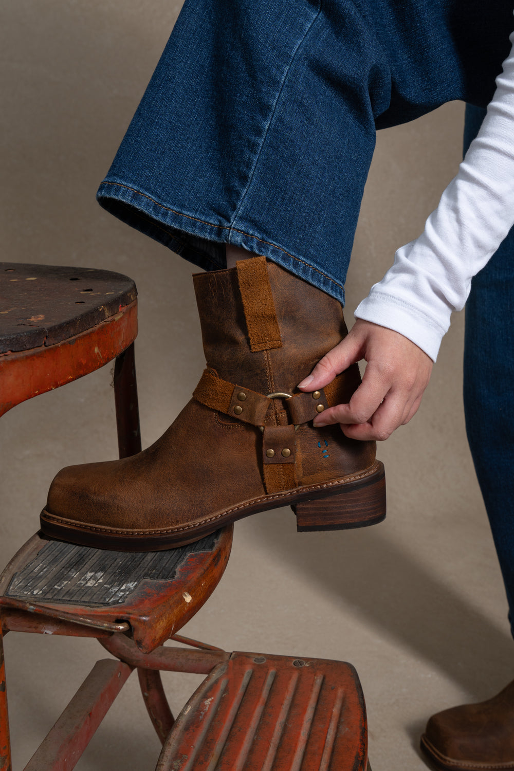 Wearing blue jeans and a white long-sleeve shirt, a person adjusts a Roan Bonsai brown full-grain leather boot while resting their foot on a worn red metal step stool.