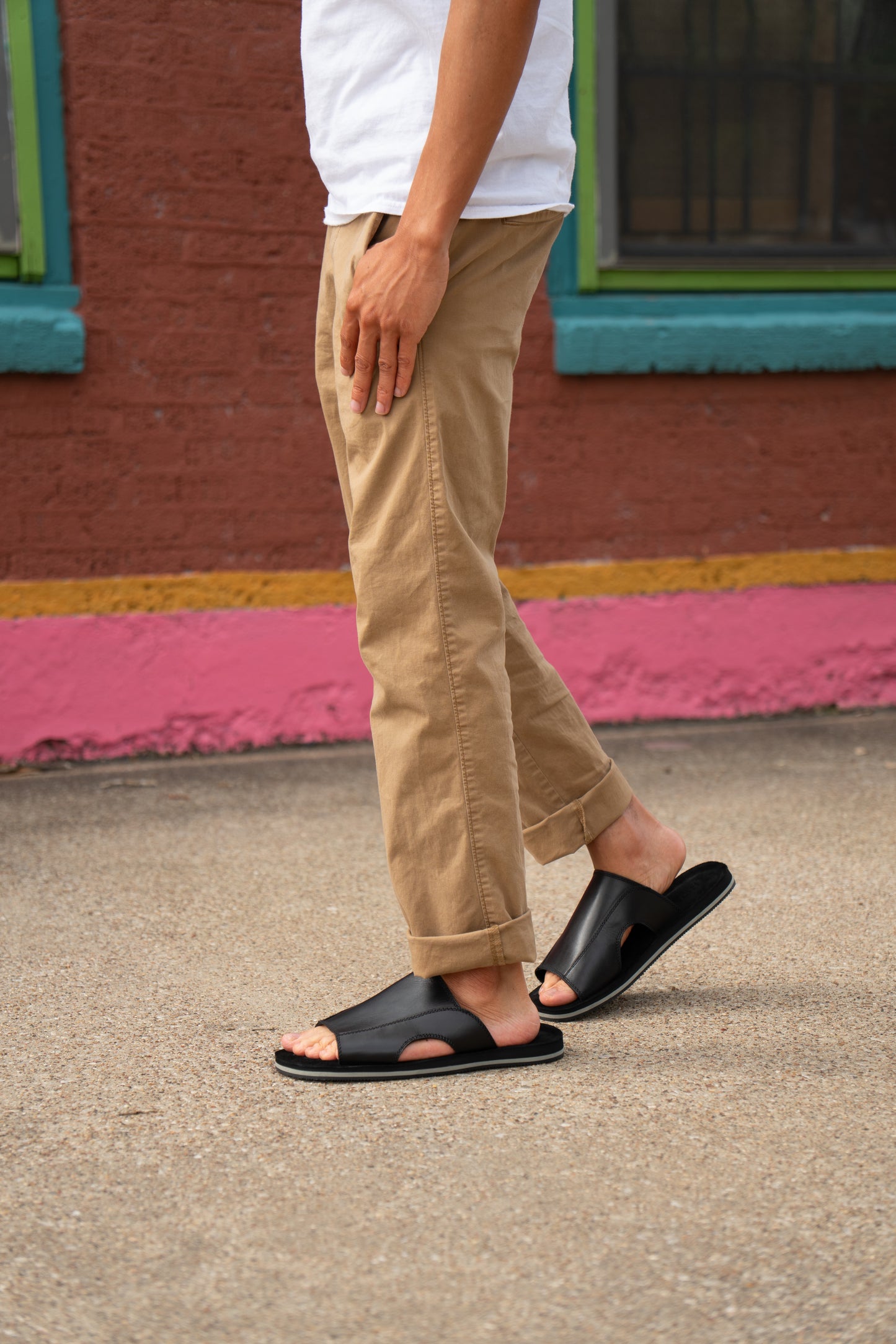 
                  
                    A person wears Roan's Breakfast tan pants and white shirt with comfortable black slip-on sandals, walking on a paved surface in front of a building with colorful trim.
                  
                