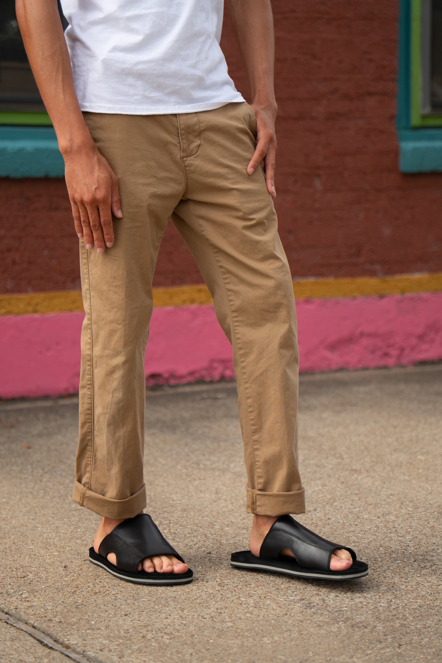A person wearing Roan's Breakfast slip-on sandals in black stands on a sidewalk in front of a brick wall with colorful trim, dressed in khaki pants and a white shirt.