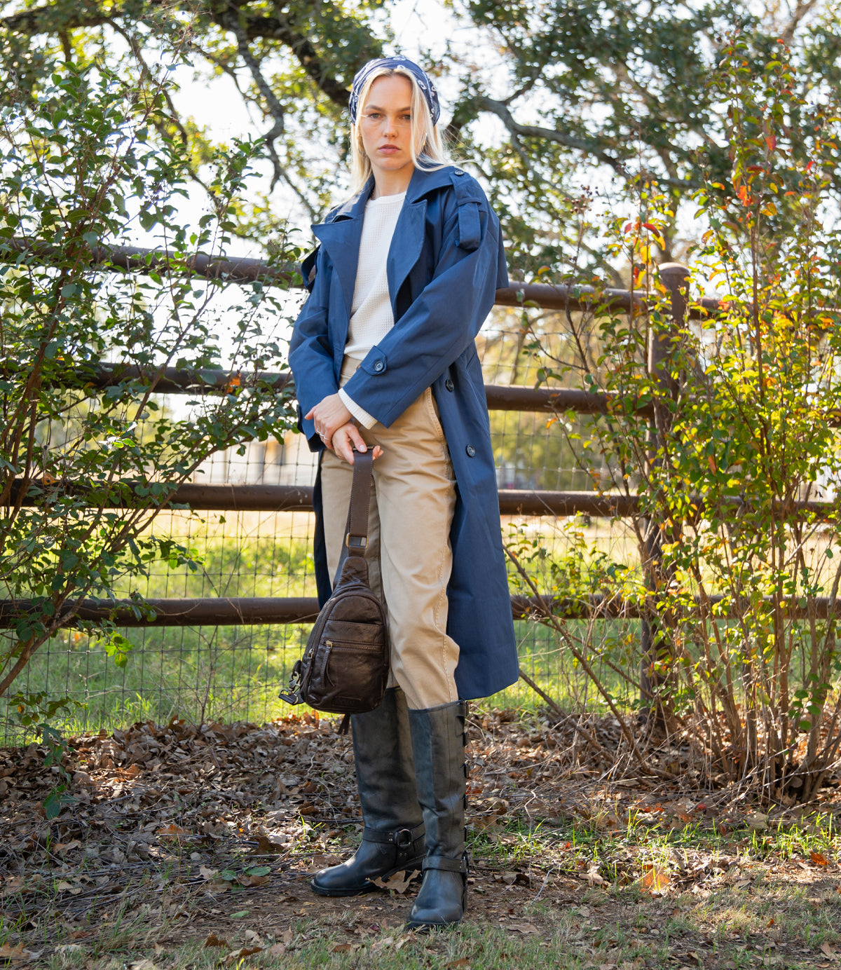 
                  
                    A woman stands outdoors by a fence, wearing Roan’s Trickle blue trench coat with a white sweater, beige pants, black boots, and carrying a compact leather bag. Trees and foliage create a lush backdrop for this versatile look.
                  
                