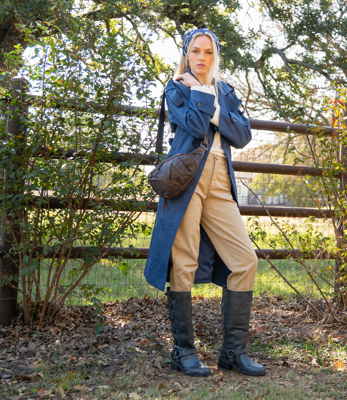
                  
                    A woman wearing a blue trench coat, tan pants, and black boots stands by a wooden fence outdoors, holding the Roan Trickle compact leather bag and looking at the camera.
                  
                