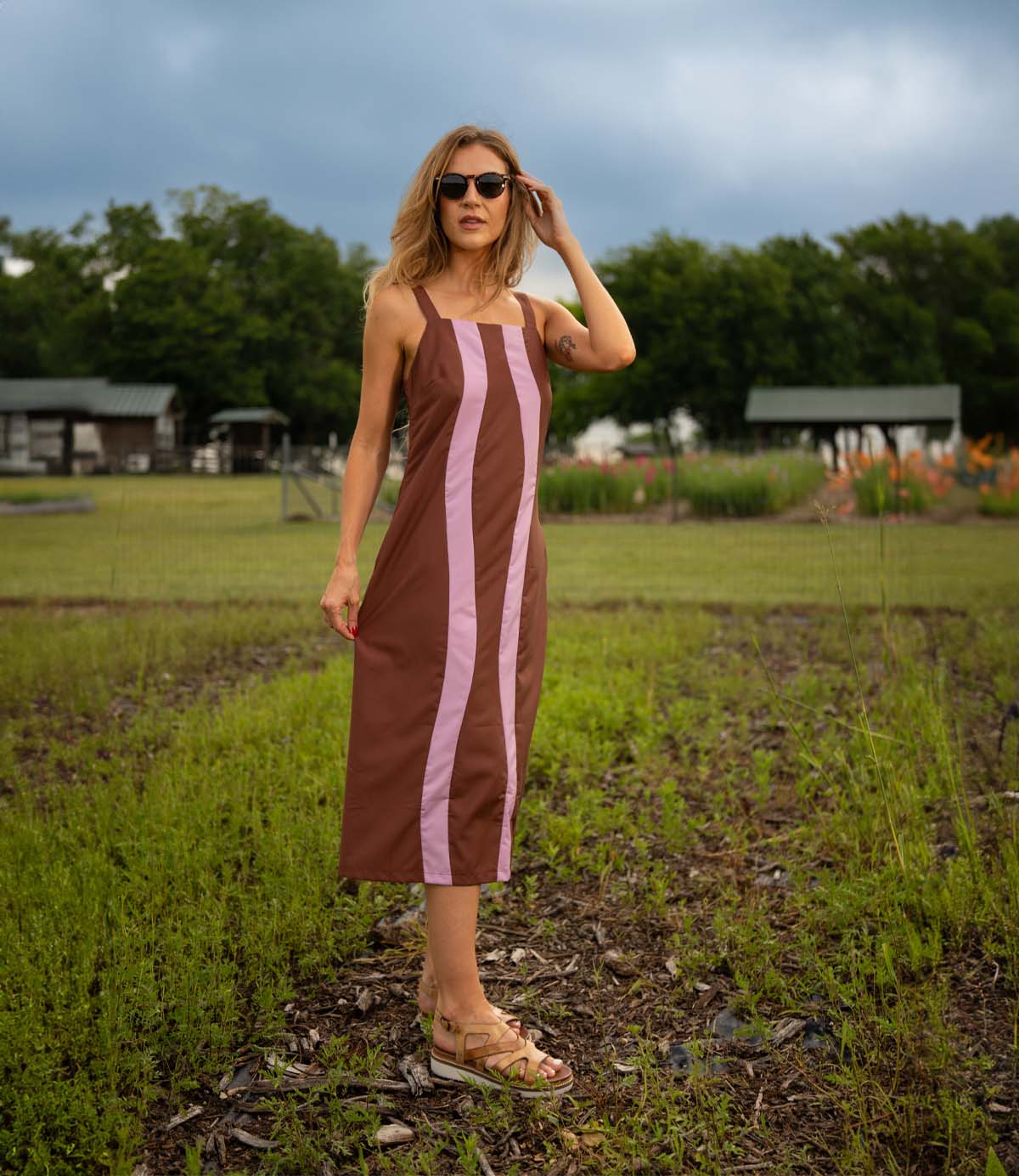 
                  
                    A woman in sunglasses and a brown dress with pink stripes stands on a grassy field, stylishly wearing Roan’s Centrist fisherman sandals with a leather upper. Buildings and trees are visible in the background.
                  
                