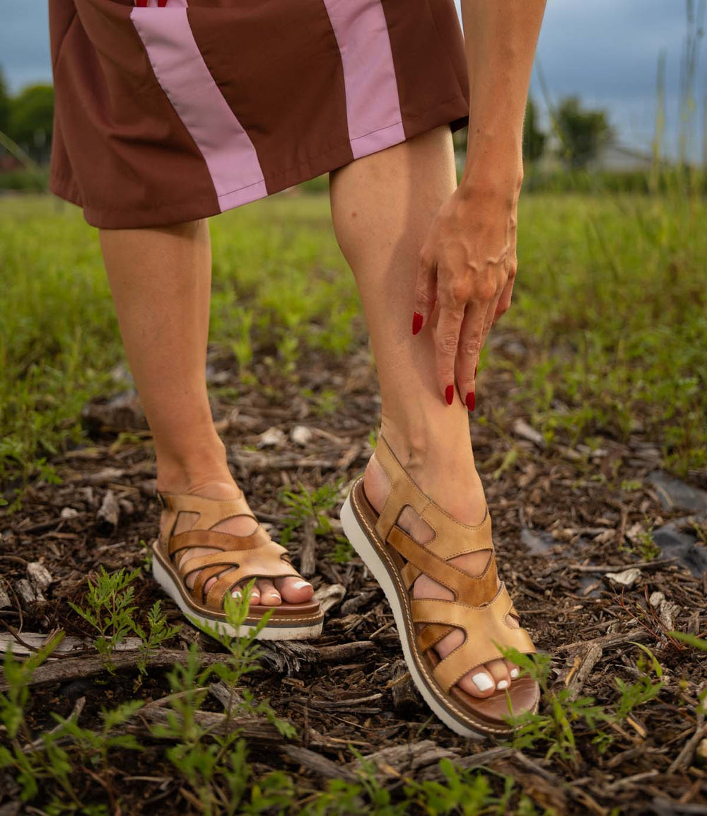 A person stands outdoors in a grassy, mulched area, touching their ankle while wearing Roan Centrist brown fisherman sandals with a leather upper and a brown skirt with pink stripes.