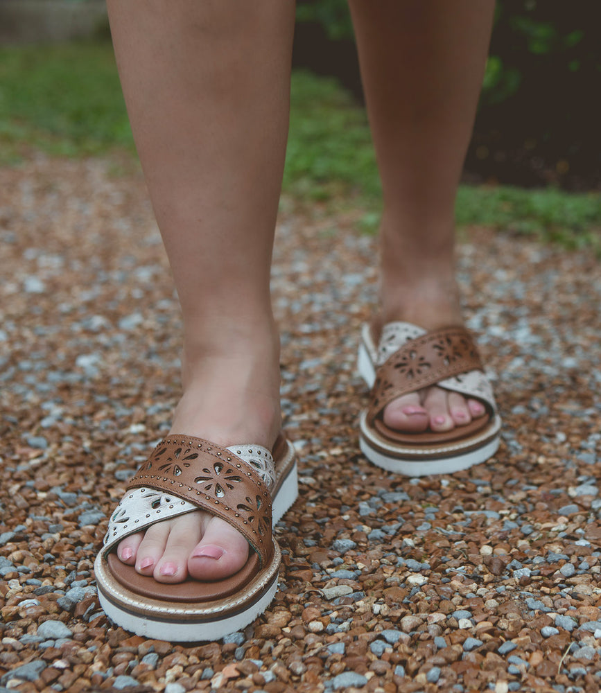 
                  
                    A person wearing Roan Chant sandals with cut-out floral patterns and leather cross straps stands on a gravel path.
                  
                