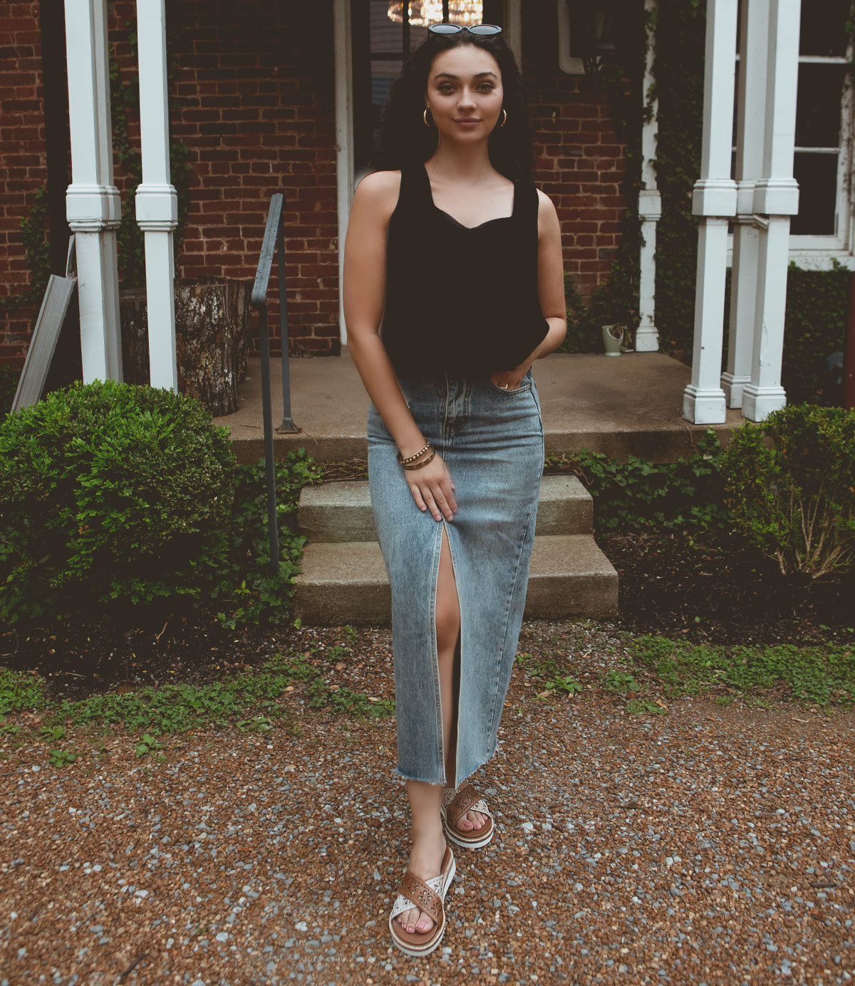 
                  
                    A woman stands in front of a brick building, wearing a black top and a long denim skirt with a front slit. She is also wearing Roan Chant lightweight sandals with leather cross straps and has one hand in her pocket.
                  
                
