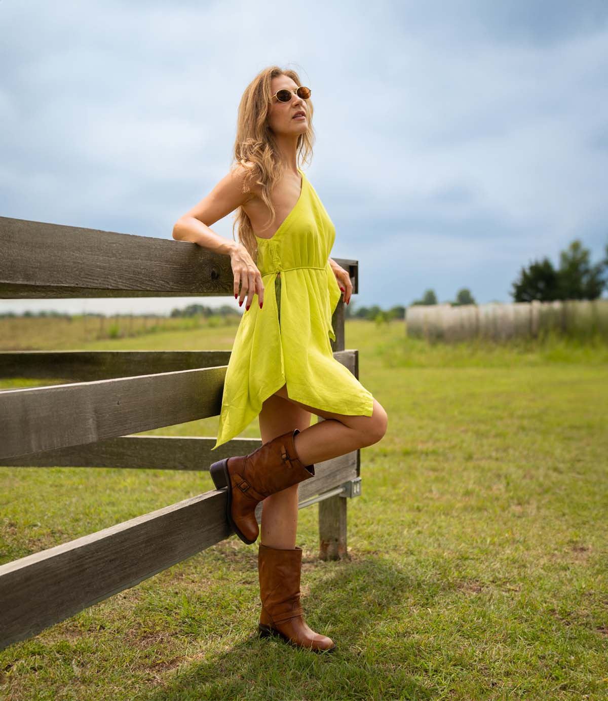 
                  
                    A woman in a yellow dress and Roan's Cite brown boots with a leather upper leans against a wooden fence in a grassy field under a cloudy sky.
                  
                
