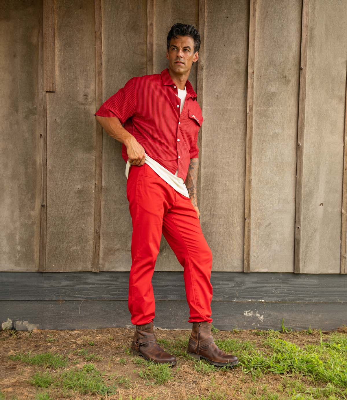 
                  
                    A man wearing Roan's Colton II hand-finished leather boots with a durable sole, a red short-sleeve shirt, red pants, and a white undershirt stands on grass against a wooden wall.
                  
                
