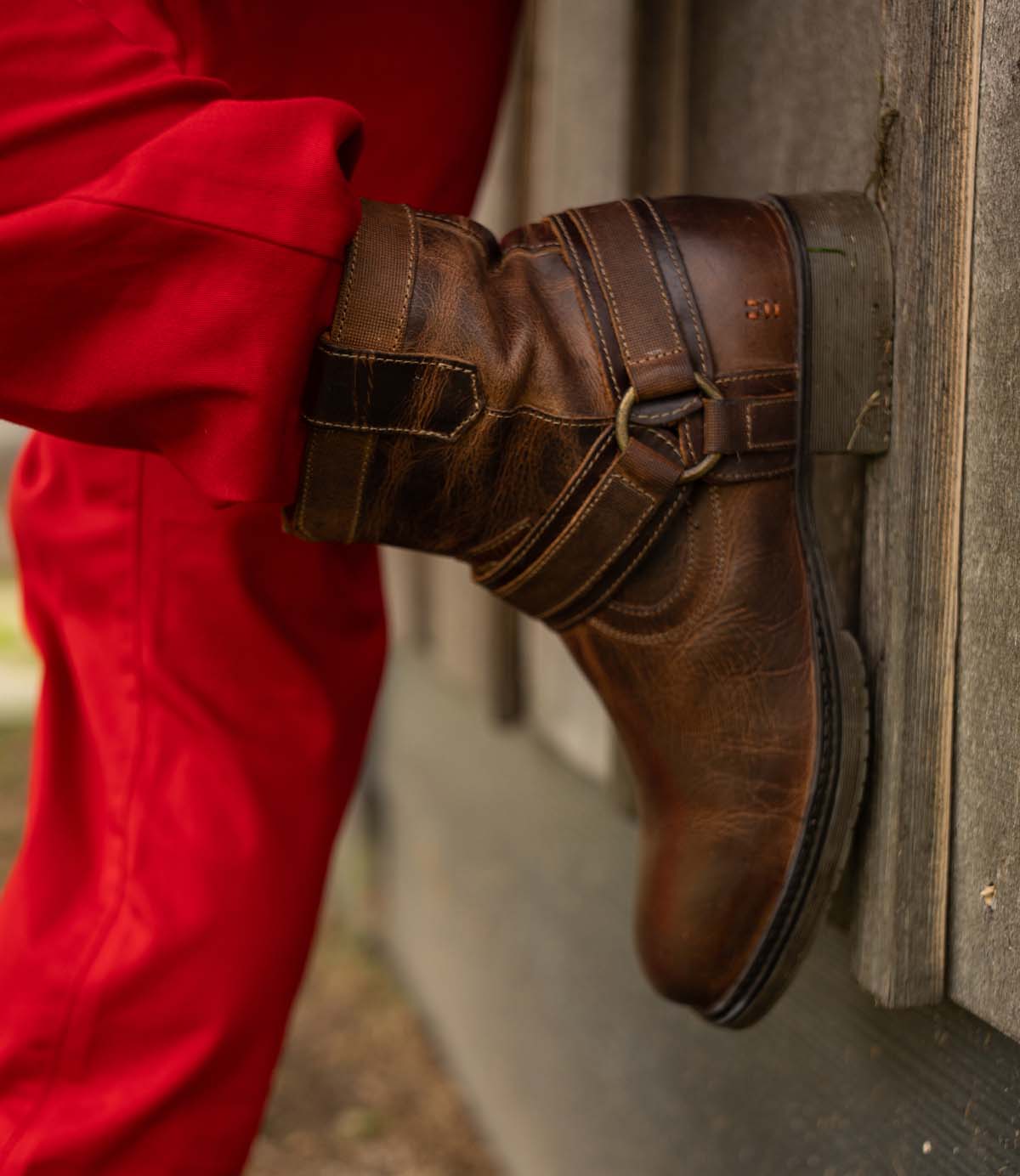 Wearing Roan’s Colton II boots—with hand-finished leather, straps, a low heel, and a durable sole—a person in red pants leans their leg against a wooden wall.
