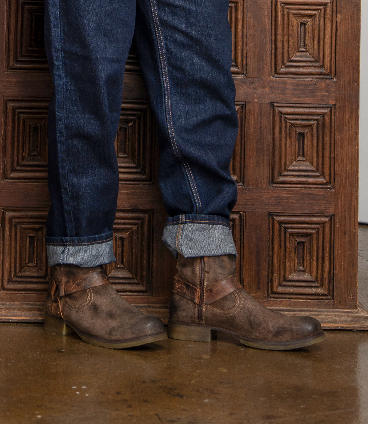 Wearing cuffed dark blue jeans and Roan’s Colton V hand-finished boots, a person stands on a polished concrete floor before a wooden paneled wall.