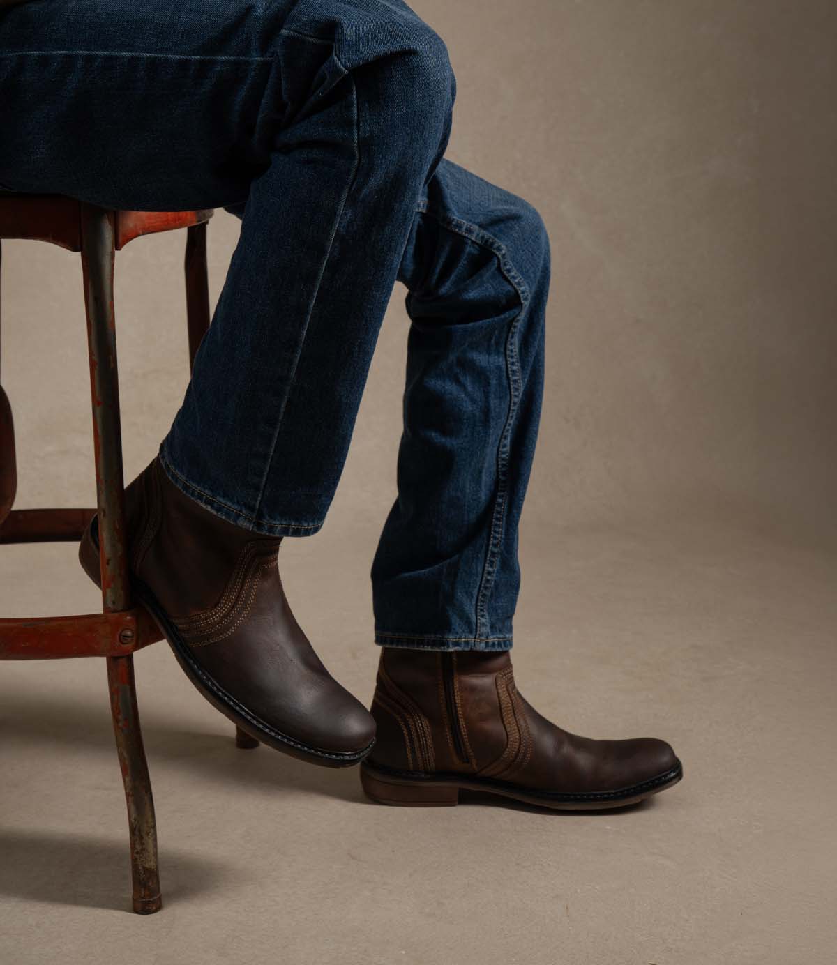 
                  
                    A person wearing blue jeans and the Roan Crestone boot, known for its comfortable leather design and sturdy rubber sole, sits on a wooden chair against a plain background.
                  
                