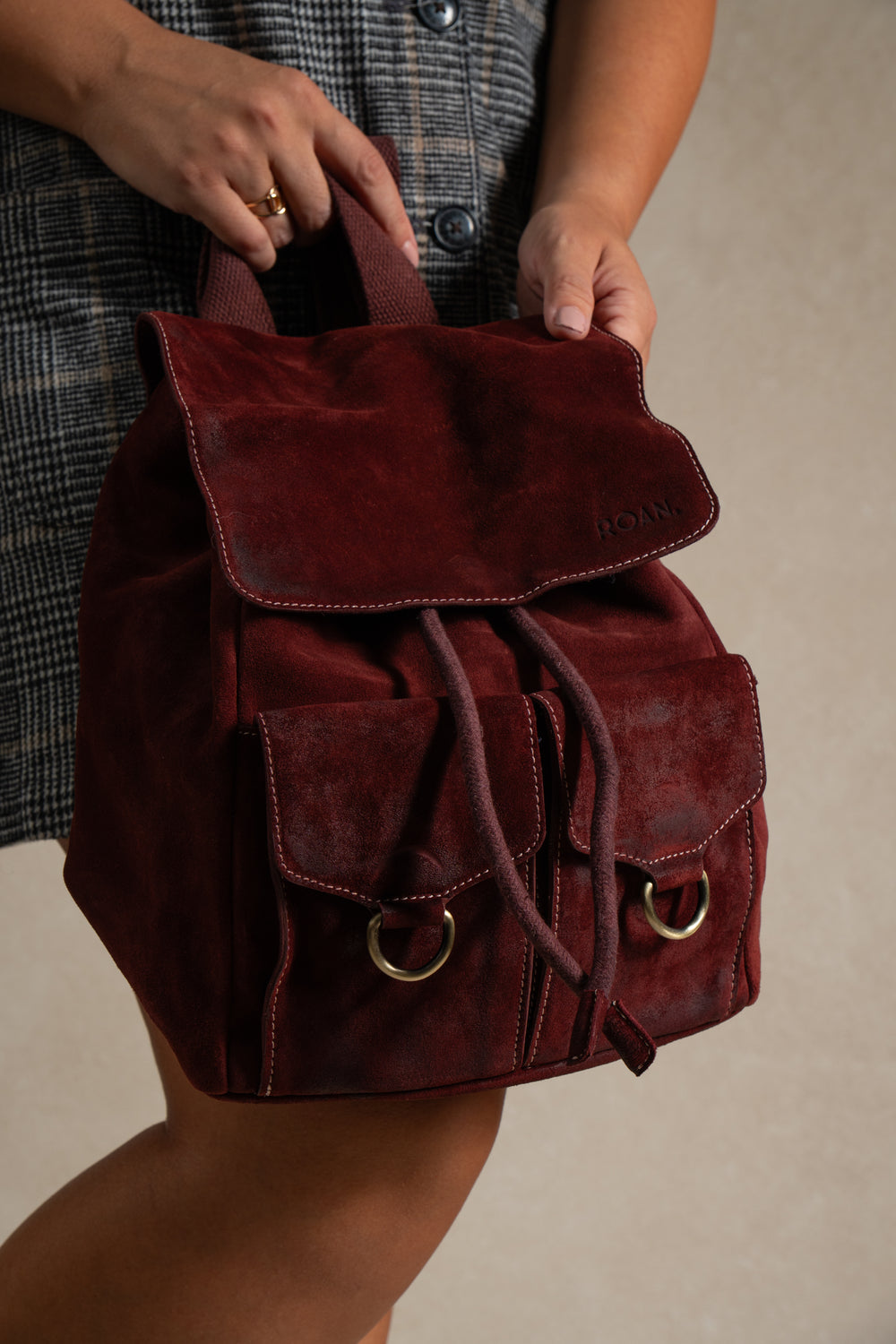 A person holding the Roan Damstad II, a premium burgundy suede backpack with two front pockets and a drawstring closure, featuring adjustable cotton straps, paired with a plaid buttoned dress.