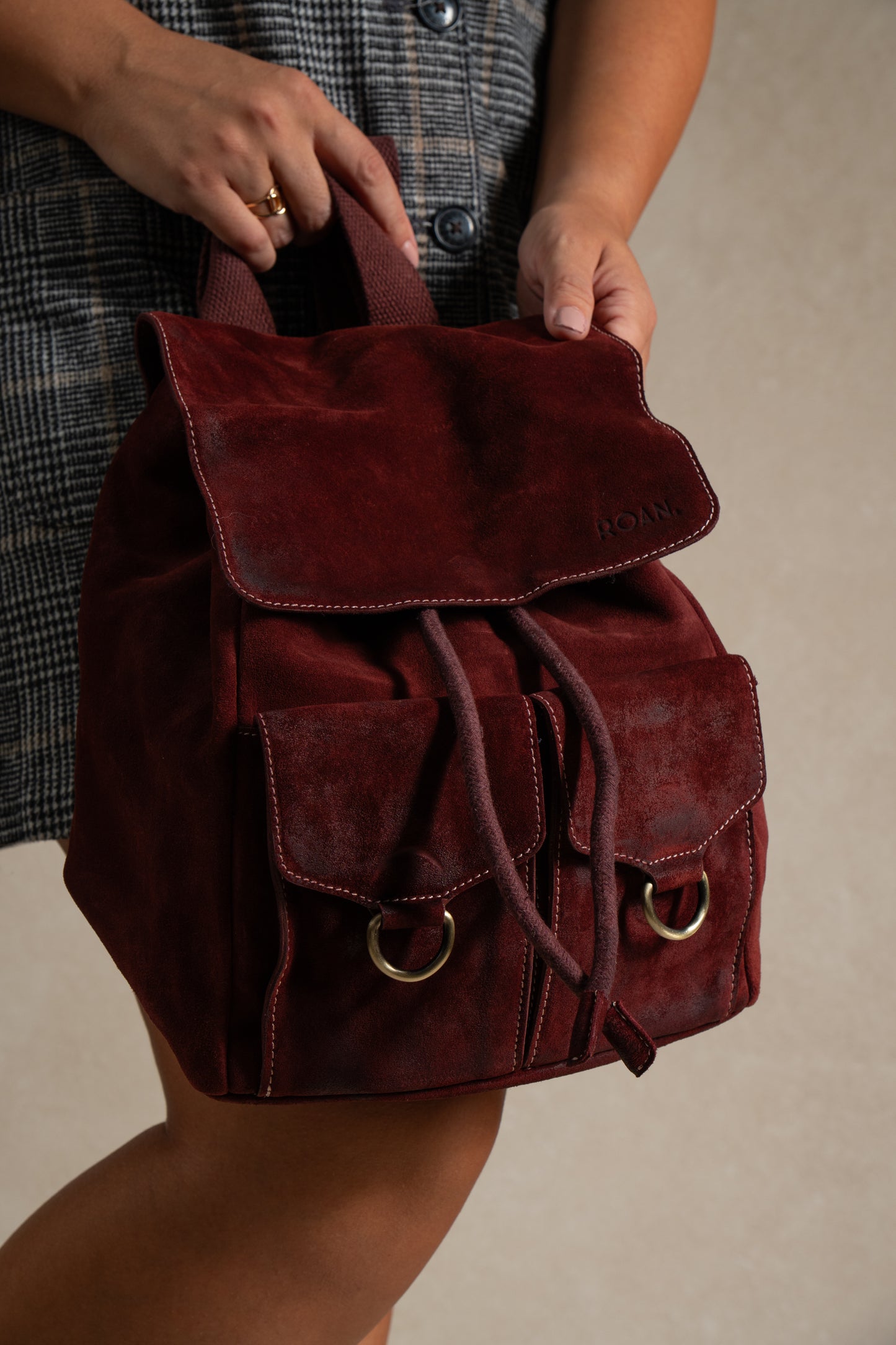 
                  
                    A person holding the Roan Damstad II, a premium burgundy suede backpack with two front pockets and a drawstring closure, featuring adjustable cotton straps, paired with a plaid buttoned dress.
                  
                