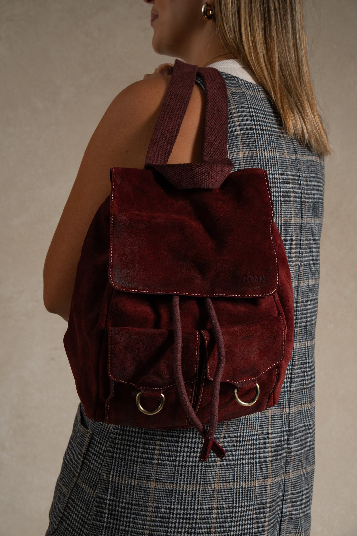 
                  
                    A woman wearing a plaid sleeveless top carries the Roan Damstad II, a premium suede backpack featuring front pockets, gold-tone hardware, and a convenient drawstring closure.
                  
                