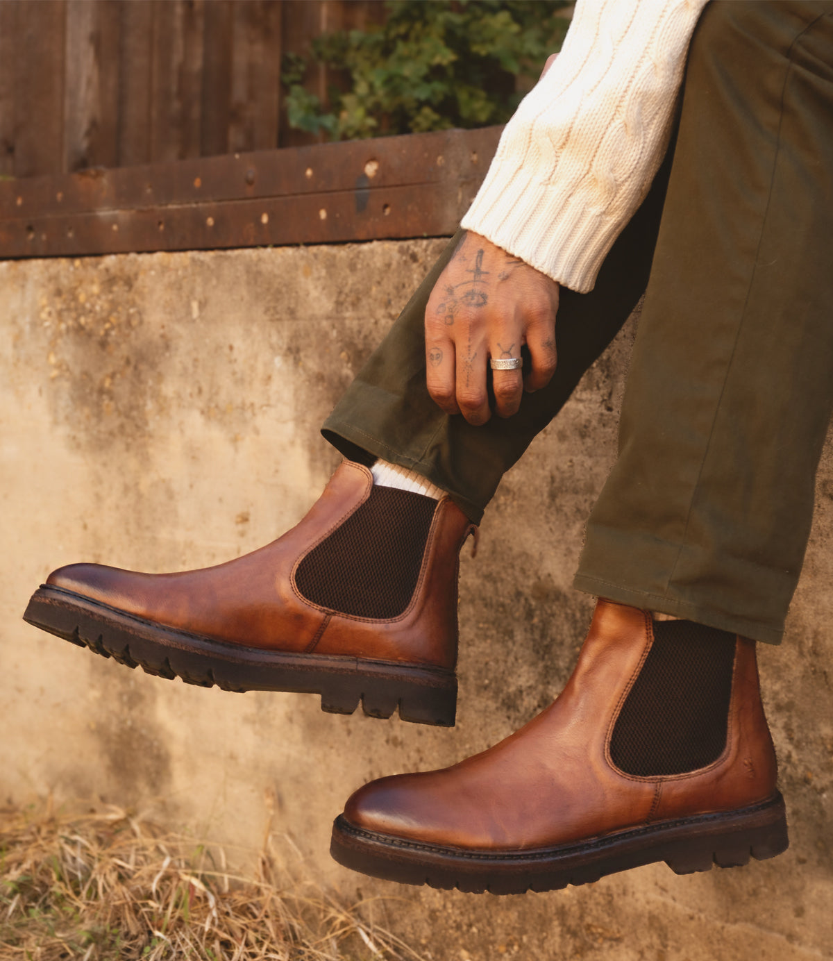 A person wearing Roan's Defy brown leather hand-finished Chelsea boots with dark elastic panels, olive pants, and a cream sweater, sitting on a stone ledge.