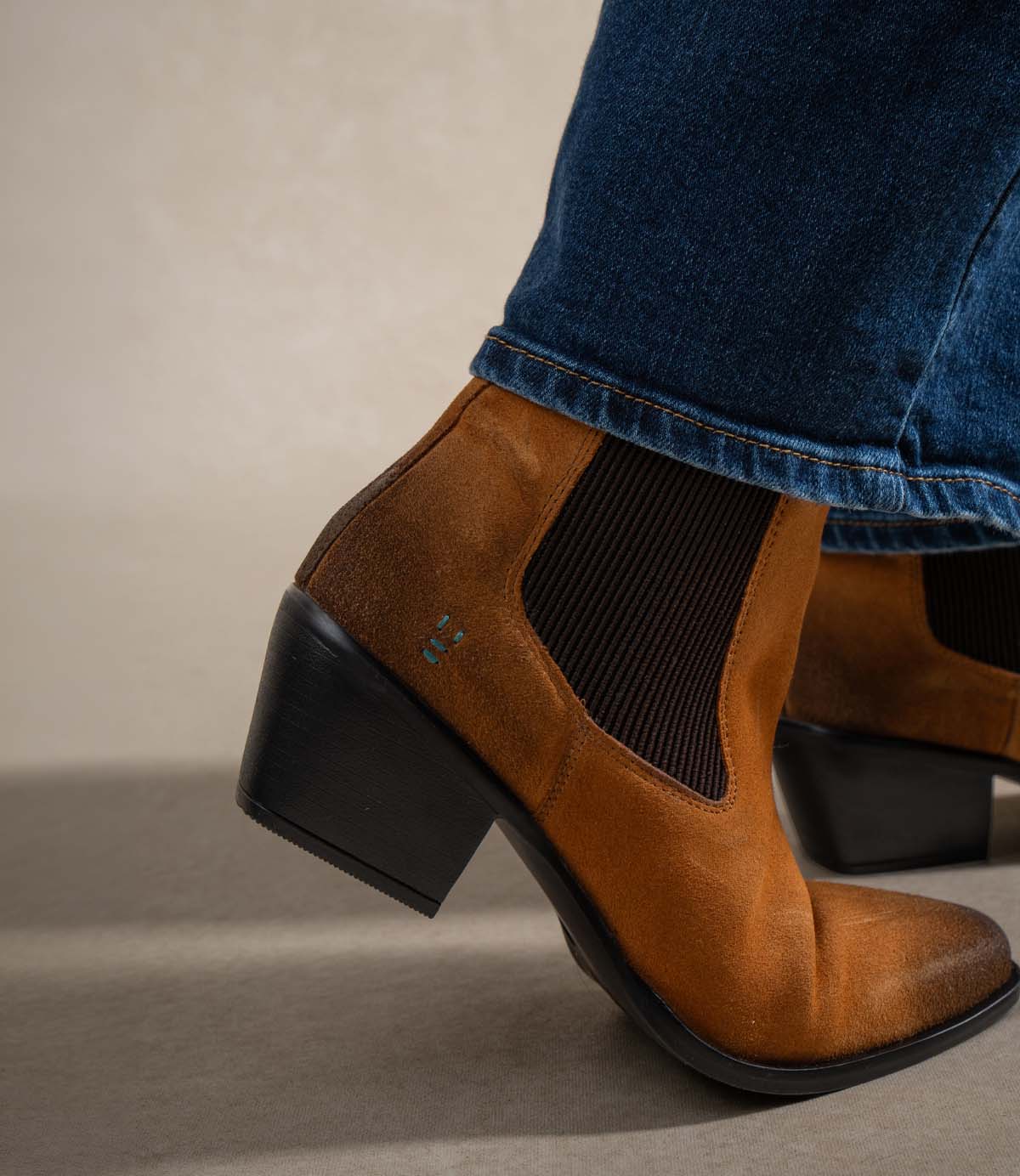 
                  
                    Close-up of a person wearing blue jeans and Roan’s Dialgoue boot—a Chelsea-inspired brown suede style with a chic block heel and stretchy side panels.
                  
                