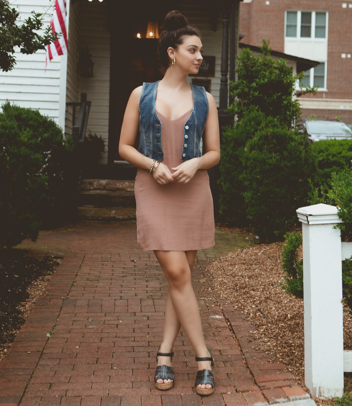 
                  
                    A woman stands on a brick pathway wearing a brown dress, denim vest, and Roan Different platform wedge heels, with her hands resting on a fence post. A building with an American flag is in the background.
                  
                