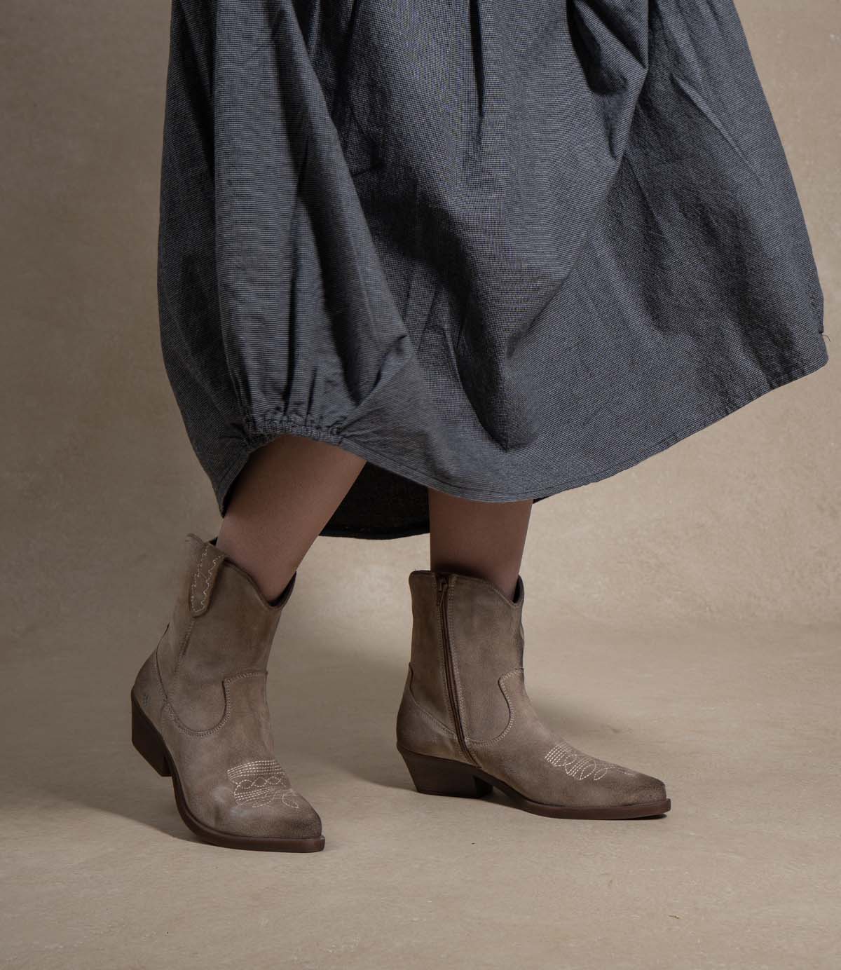 
                  
                    A person wearing Roan’s “Discussion” loose, dark gray skirt and taupe cowboy boots with a soft suede finish stands on a beige surface with legs slightly apart, channeling a confident western attitude.
                  
                