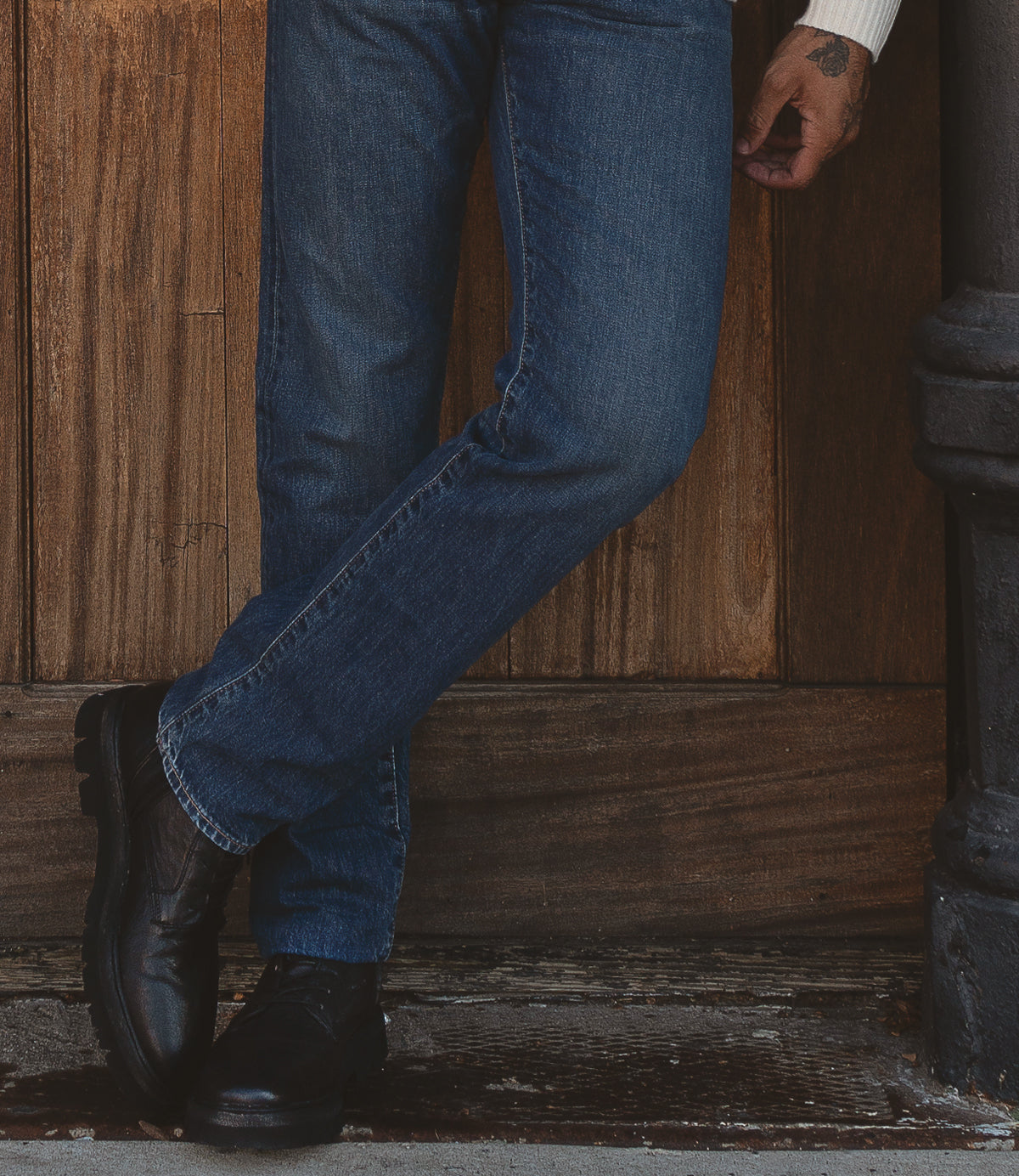 A person dressed in Roan's Diss jeans and black combat-style boots leans against a wooden wall.