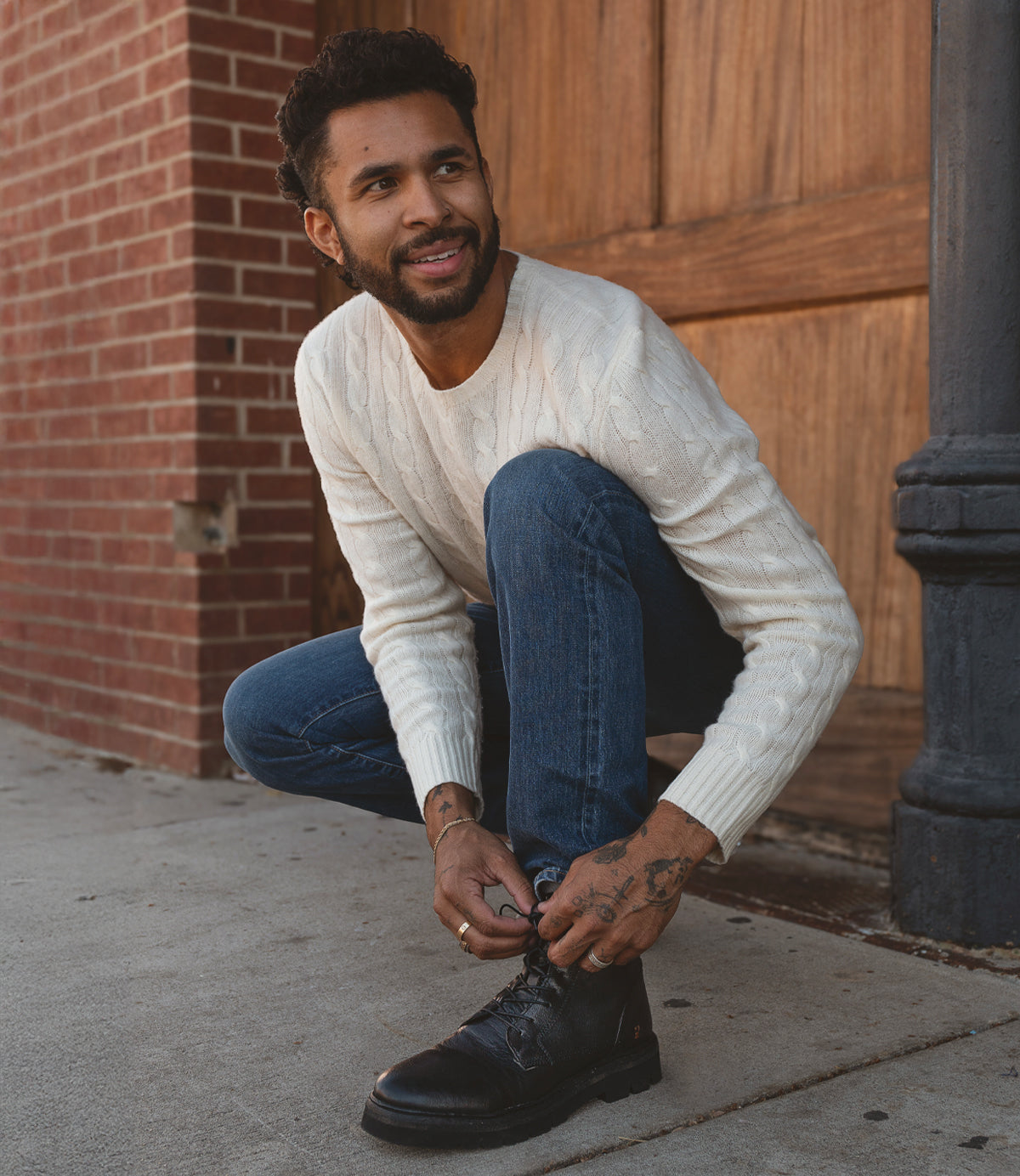 
                  
                    A man kneels on the sidewalk to tie his Roan Diss combat-style boot, known for its EVA outsole that offers comfort and durability. He wears a white sweater and blue jeans, framed by a brick and wooden background.
                  
                
