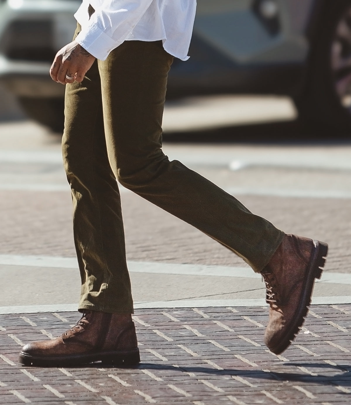 A person strides confidently across the street in Roan's Duty brown suede boots, featuring a convenient side zipper, perfectly paired with olive pants.