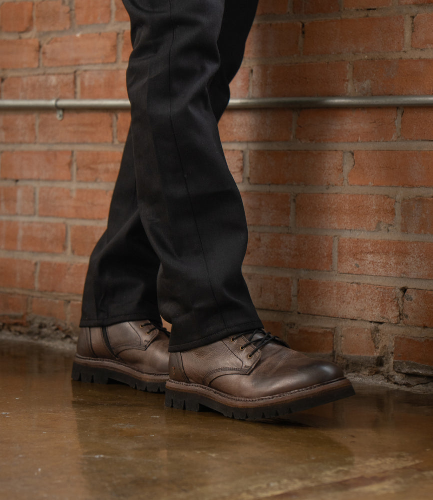 
                  
                    A person wearing black pants and Roan Duty brown leather men's boots with a padded insole stands on a polished floor against a brick wall background.
                  
                