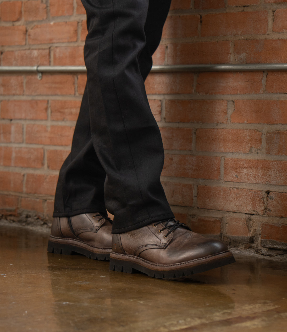 
                  
                    A person wearing black pants and Roan Duty brown leather men's boots with a padded insole stands on a polished floor against a brick wall background.
                  
                