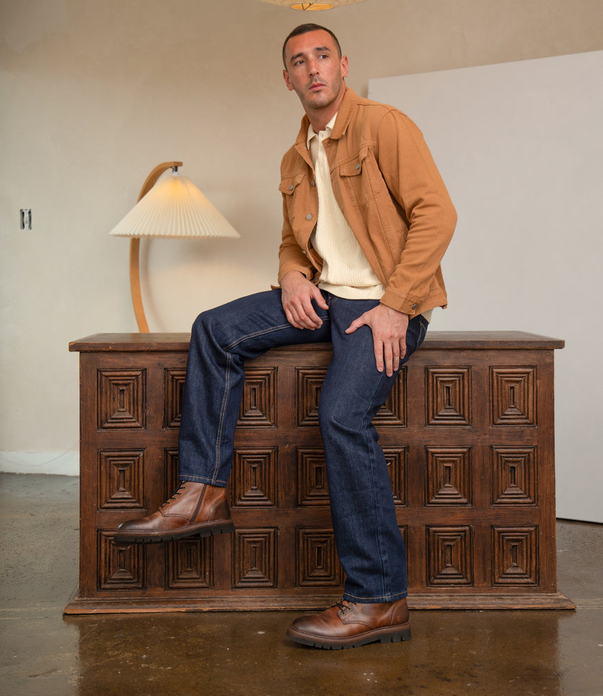 
                  
                    A man wearing Roan Duty brown suede boots with a padded insole, a tan jacket, cream shirt, and dark blue jeans sits on a carved wooden chest in a room with a lamp and light walls.
                  
                