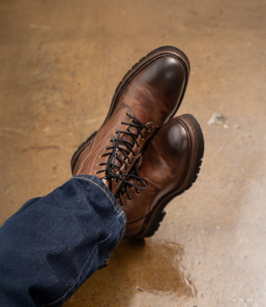 A person wearing Roan Duty dark blue jeans and brown men's boots with a padded insole sits cross-legged on a slightly reflective brown floor.