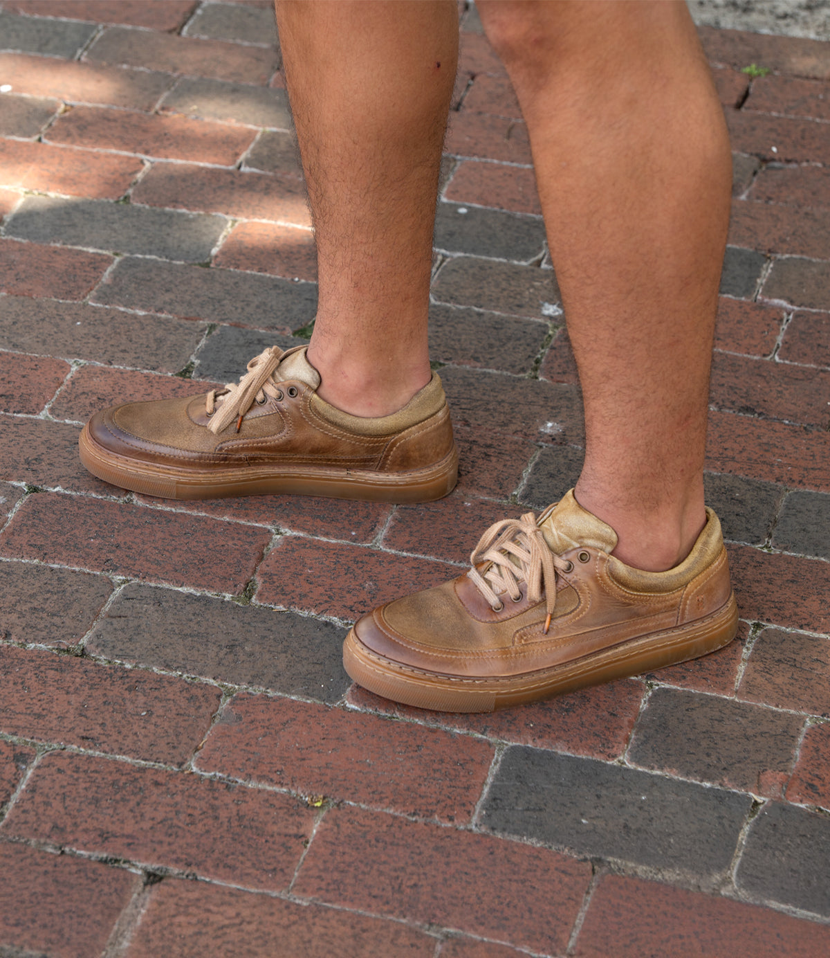 A person wearing Roan Eminent brown lace-up sneakers made of worn top-grain leather stands on a brick pavement.