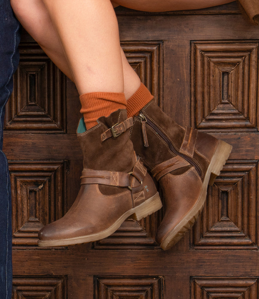 A person wears Roan’s Flair brown leather ankle boots with Western stitching and orange socks, sitting cross-legged before a wooden door with carved rectangular panels.