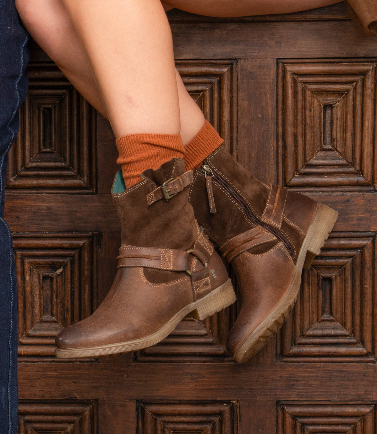 A person wears Roan’s Flair brown leather ankle boots with Western stitching and orange socks, sitting cross-legged before a wooden door with carved rectangular panels.