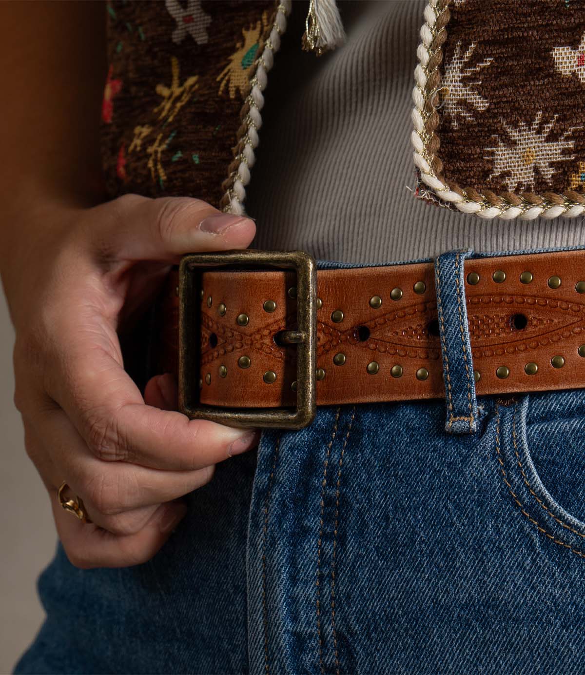 A person dressed in blue jeans, a gray shirt, and a patterned vest holds the antique brass buckle of the rugged Roan Flintrow leather belt with one hand.