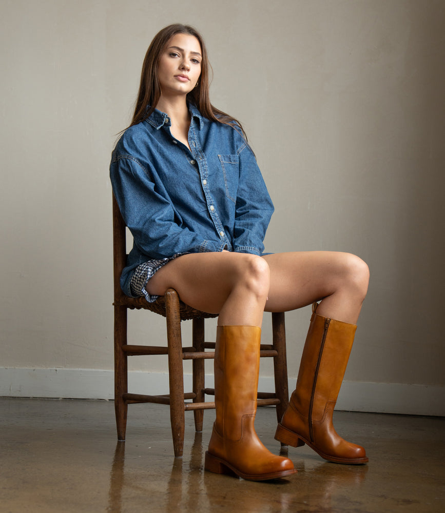 
                  
                    A woman sits on a wooden chair in a minimalist room wearing a blue denim shirt, patterned shorts, and Roan's Froe tall brown full-grain leather boots with western stitching.
                  
                