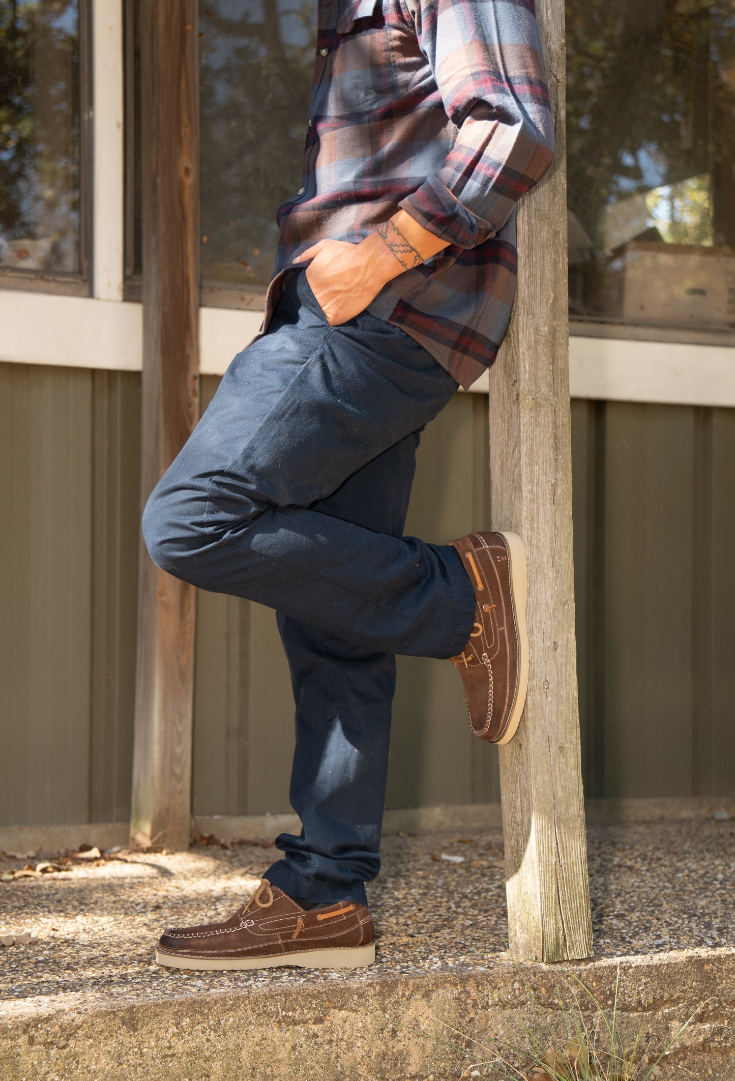 
                  
                    Wearing Roan's Glory boat shoes, a person in a plaid shirt and blue pants leans against a wooden post outdoors, one leg crossed over the other, exuding timeless nautical style.
                  
                